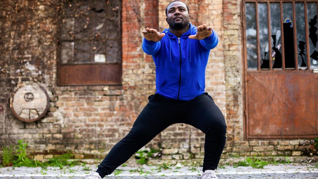 black man in blue hoodie doing side squat in front of brick wall