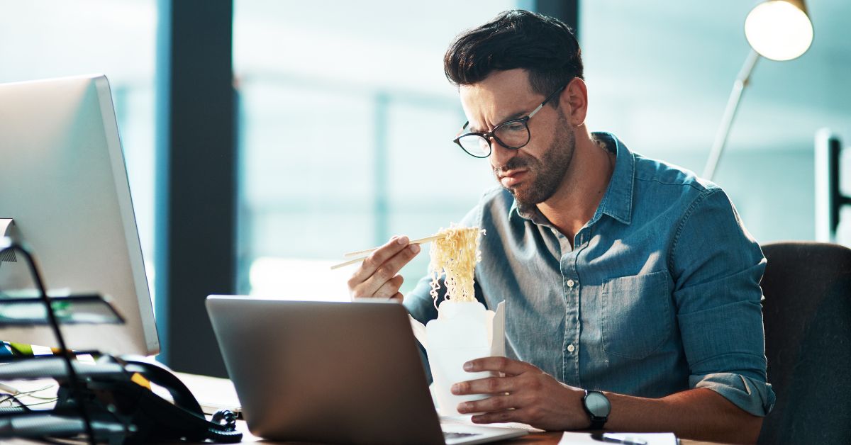 grumpy man at desk eating noodles