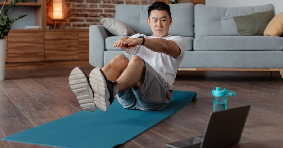 Mature korean man doing exercises for abs on mat on floor in living room interior, watching workout on laptop computer