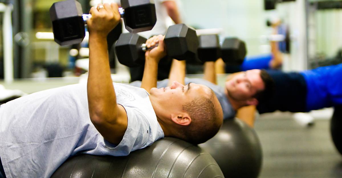 man holding dumbbell weights on a yoga ball_feat image