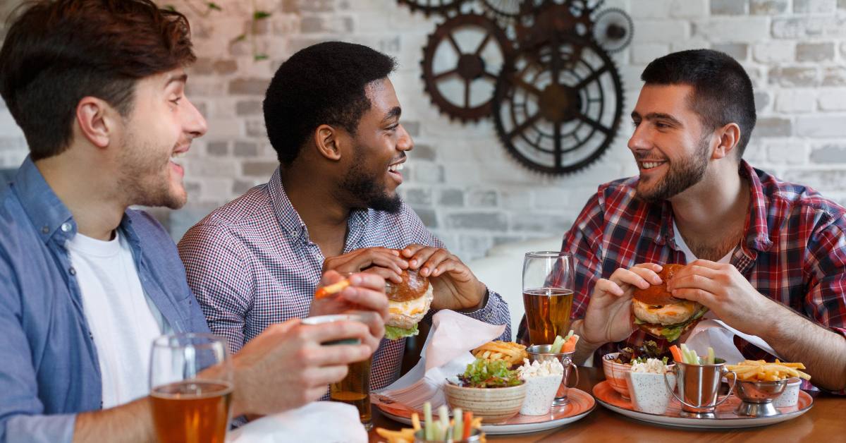 three male friends eating burgers and drinking beer in a restaurant